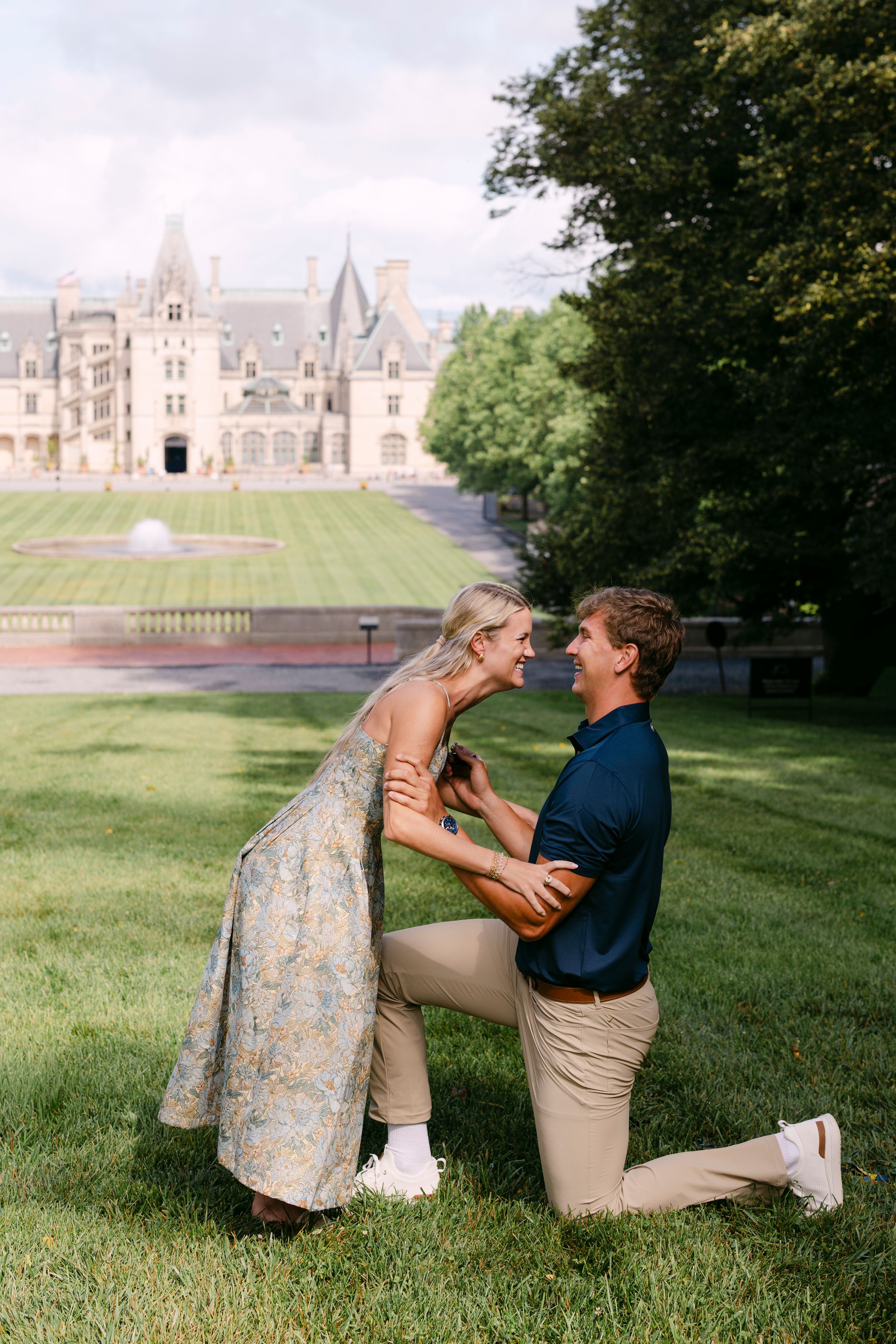 A joyful couple embraces outdoors, woman in floral dress laughing as man lifts her beneath lush green trees.