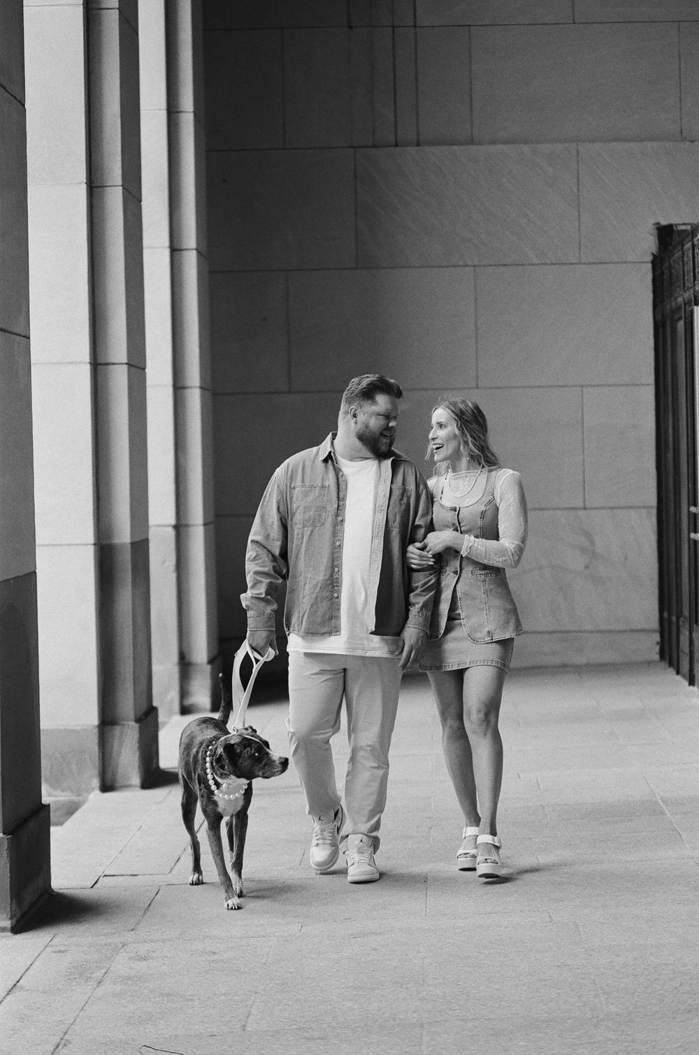Black and white photo of a couple walking their dog on a city sidewalk, smiling at each other.