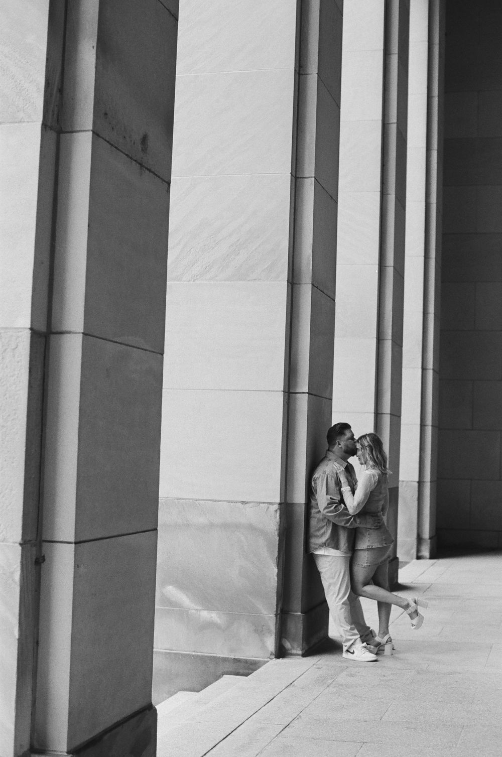 Black and white photo of a romantic couple embracing against a marble column wall outdoors.