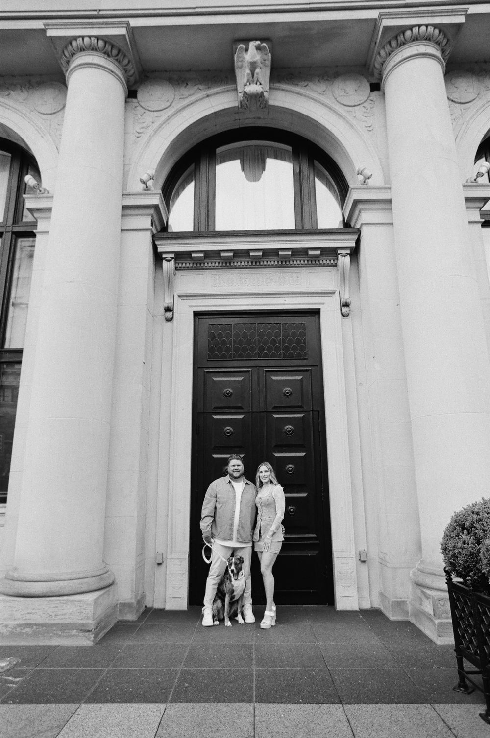 Couple stands with their dog in front of elegant neoclassical building entrance with arched doorway.