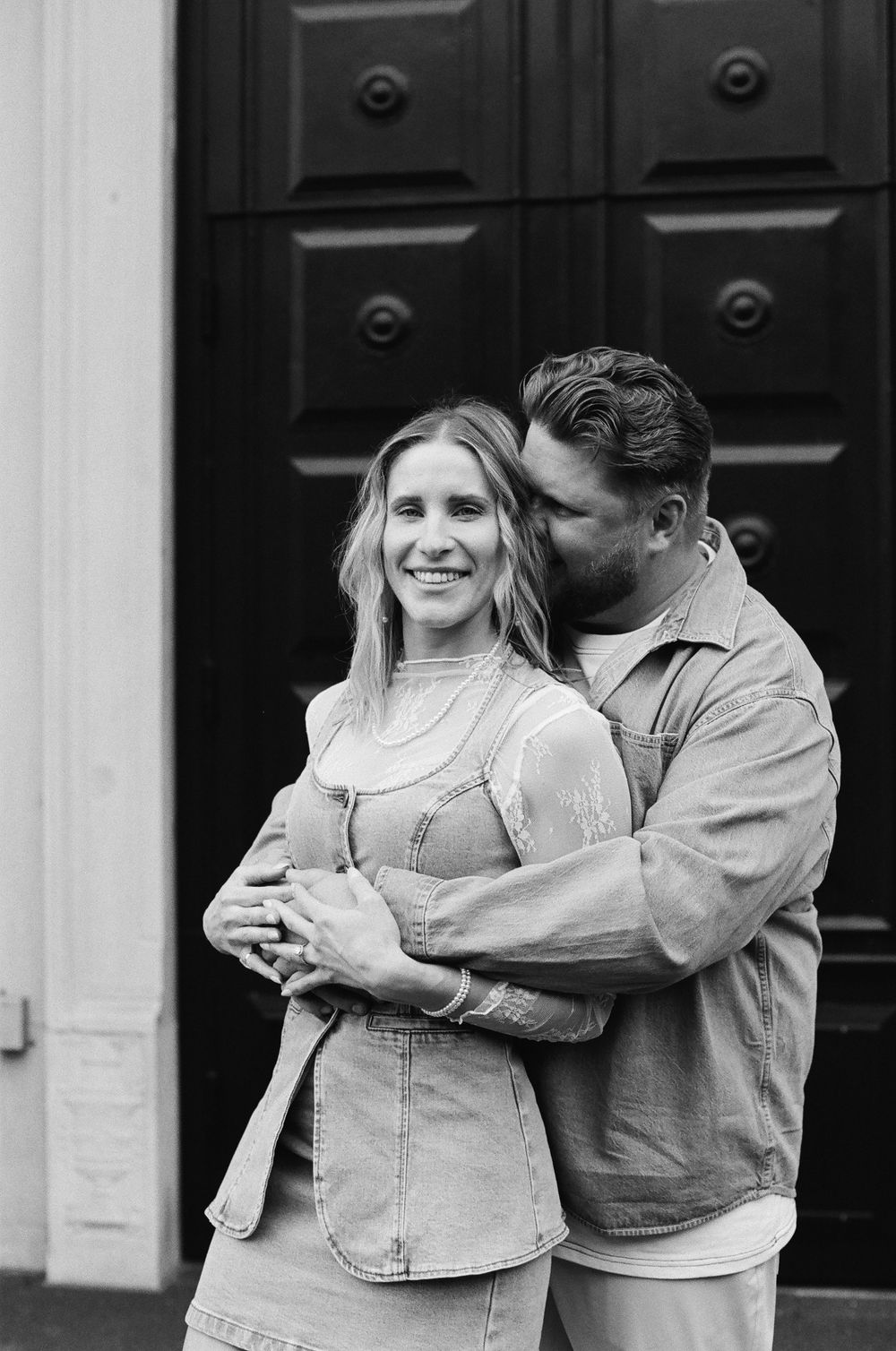 Black and white photo of a smiling couple embracing in front of a dark door, woman wearing denim dress.