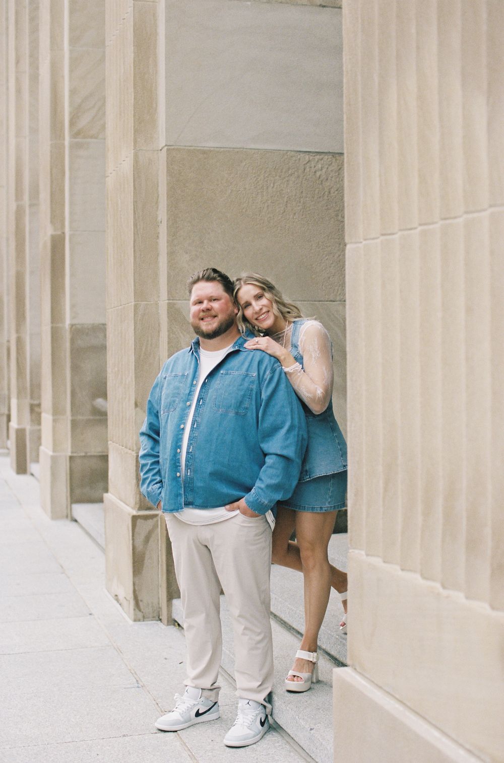 A couple poses by stone columns, man in denim shirt and khakis, woman in denim dress leaning on his shoulder.