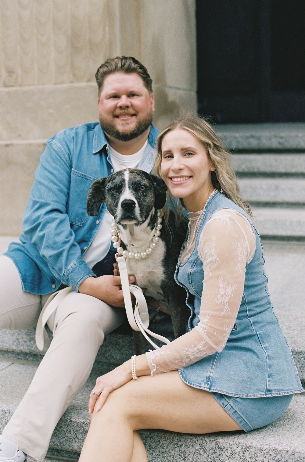 Couple sitting on stone steps with their dog wearing a pearl necklace, both dressed in denim outfits.