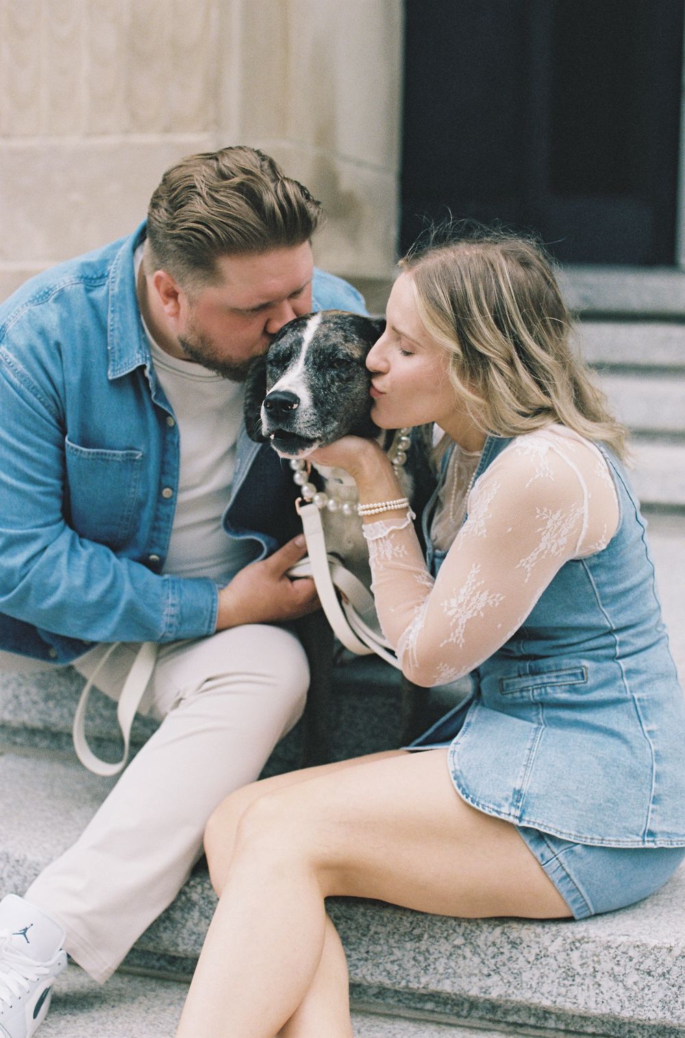 Couple sitting on stone steps kissing their black and white dog during outdoor photo session.