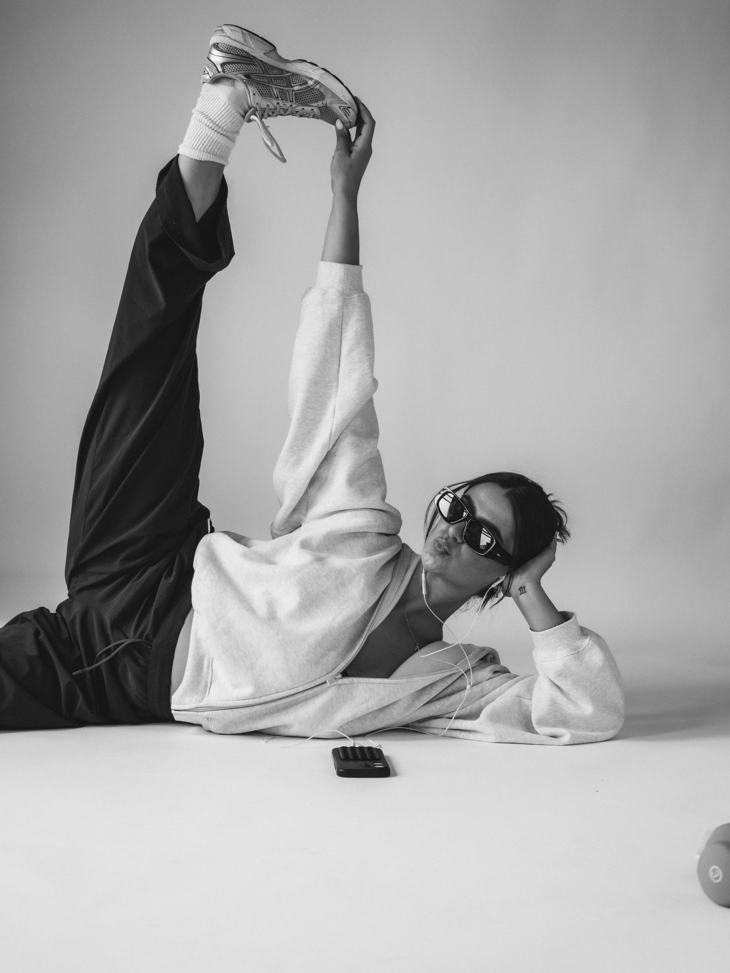 Person lying on floor in white sweatshirt and sunglasses, leg raised high wearing silver sneakers, black and white photo.