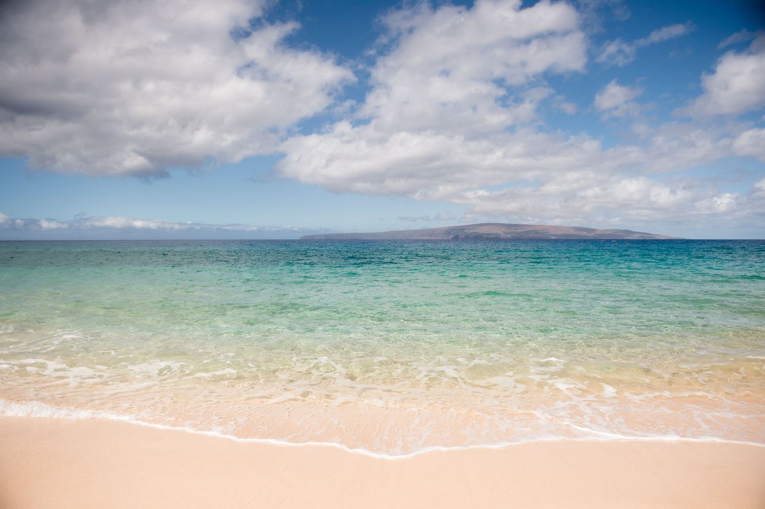 Crystal clear turquoise water lapping on a pristine white sandy beach with island in distance.