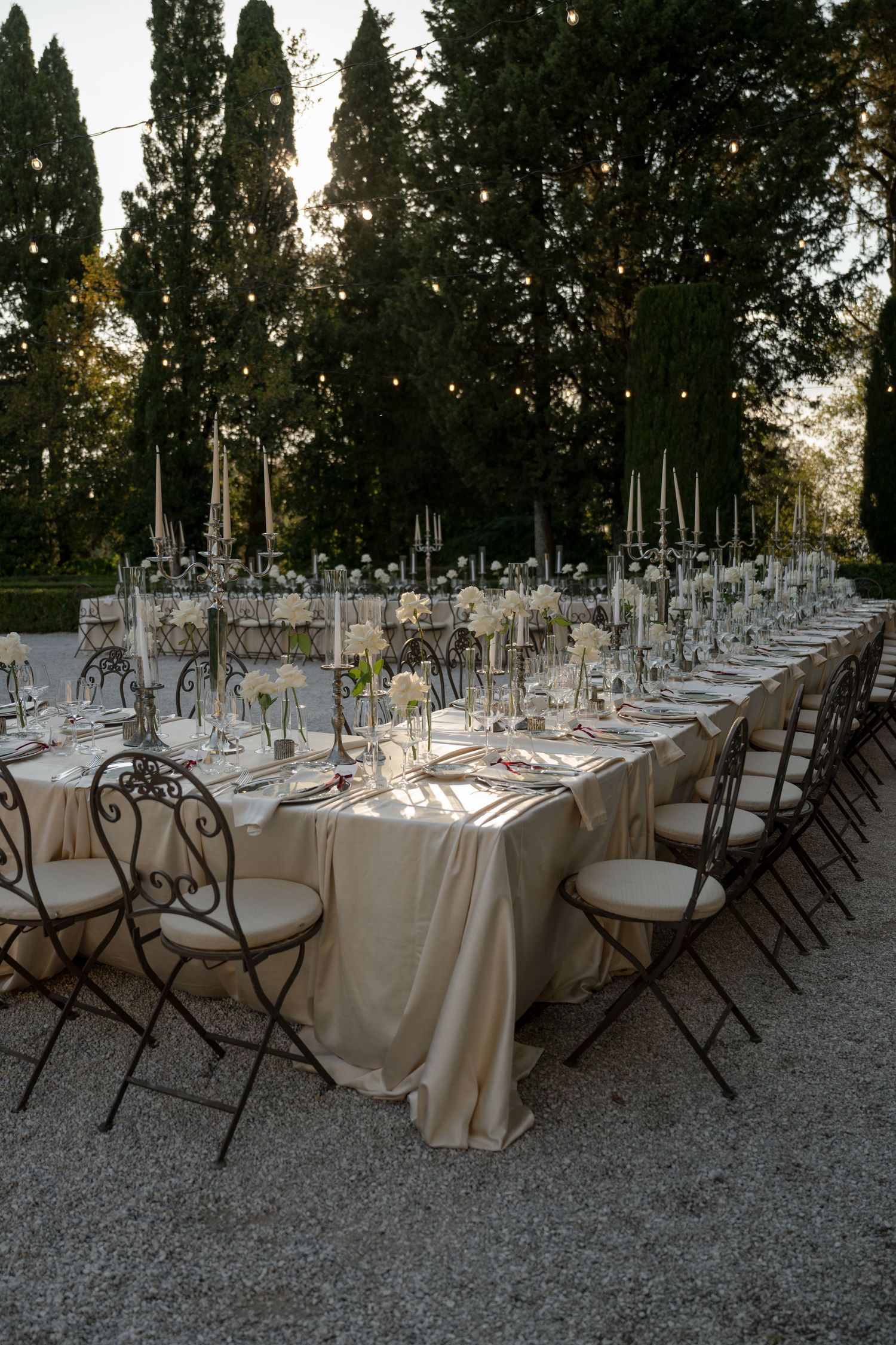Elegant outdoor wedding reception with long tables, candelabras, white florals, string lights, and tall cypress trees at dusk.