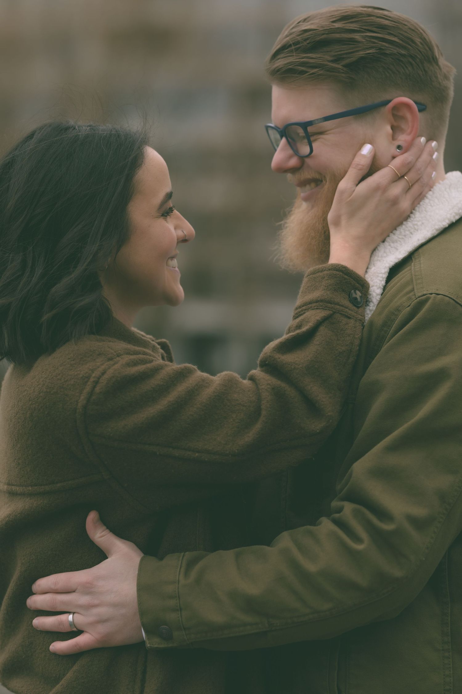 Smiling couple in olive jackets embrace and gaze at each other tenderly in an outdoor portrait.