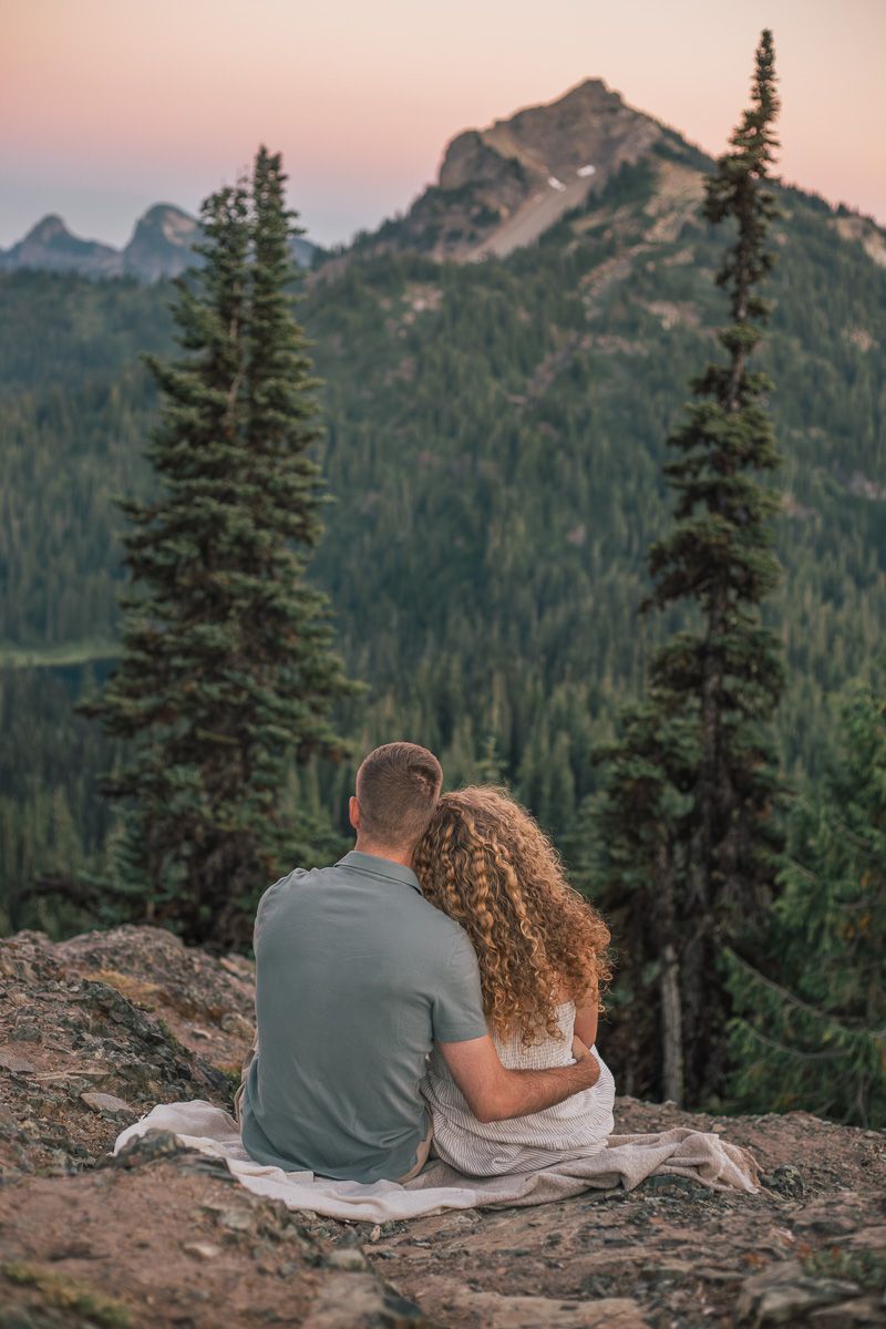 Couple sitting together on a rocky overlook enjoying a scenic mountain sunset view surrounded by tall pine trees.