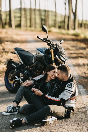 A romantic motorcycle photoshoot in a wooded area featuring a couple laying on the ground near their bike.