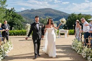 A wedding ceremony takes place on a scenic hilltop venue with white floral decorations lining the aisle.