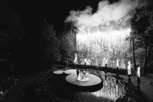 Dramatic black and white nighttime scene of people walking through a misty park with glowing lights and trees.