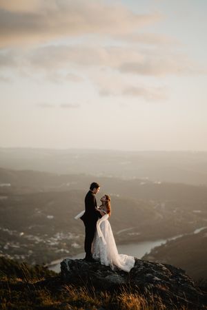 A couple in wedding attire stands silhouetted on a mountaintop overlooking a misty valley at sunset.