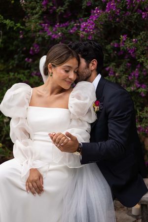 Romantic couple in wedding attire poses against purple flowering bushes.