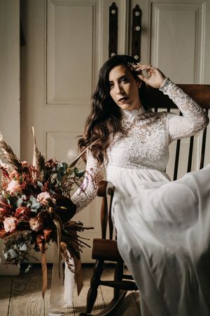 Elegant bridal portrait featuring a long-sleeved lace wedding dress and dramatic floral arrangement in moody lighting.