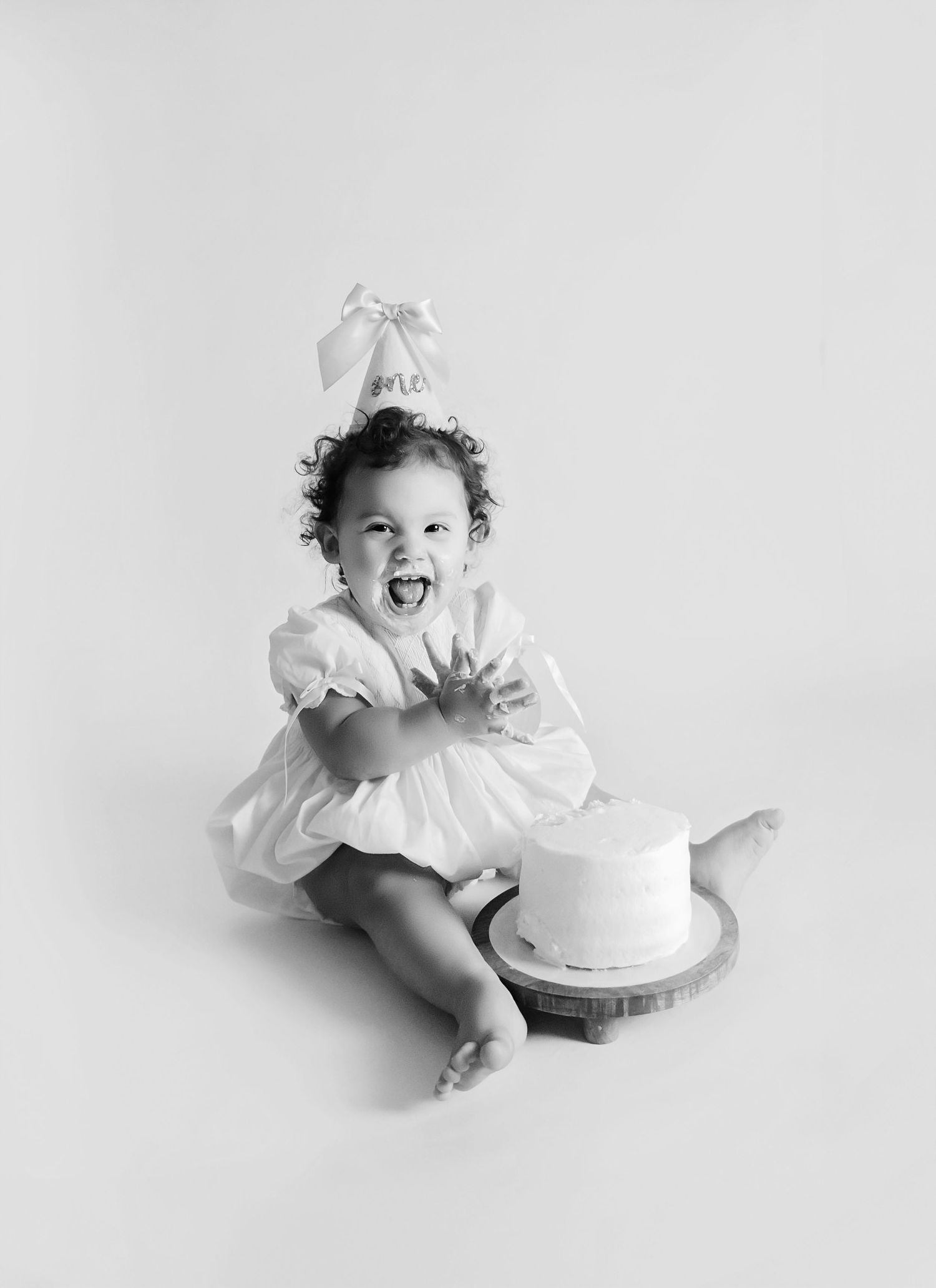 Black and white photo of a joyful baby girl in white dress celebrating first birthday smash cake session.