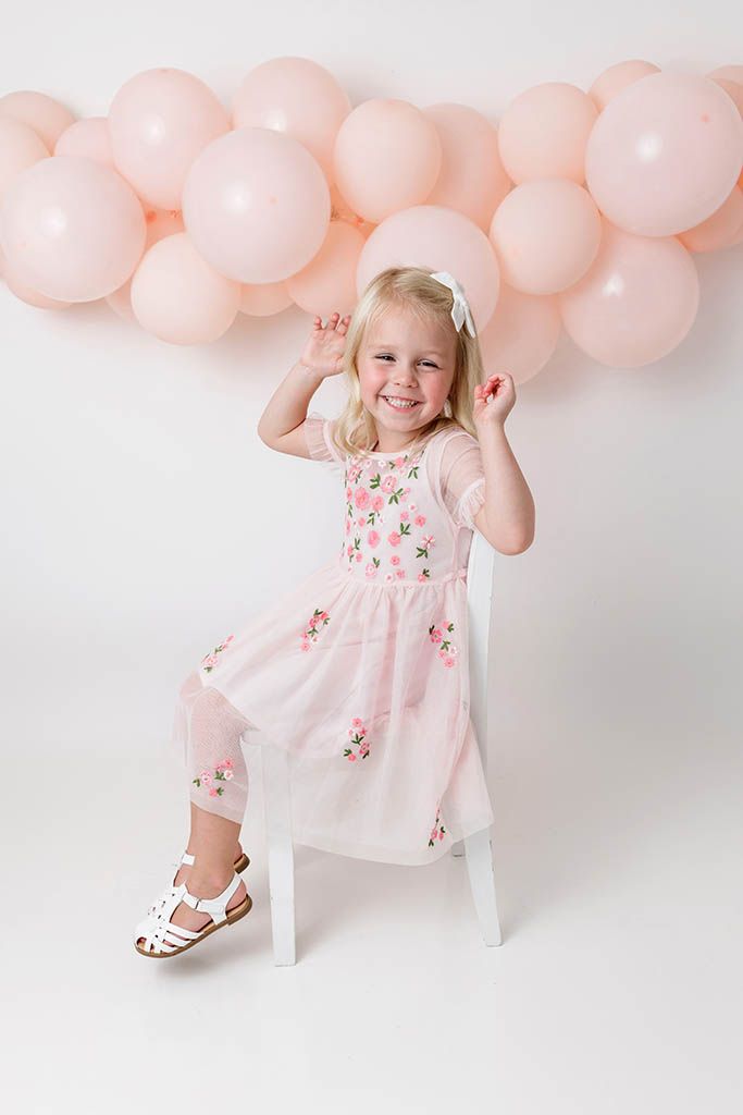 Smiling blonde girl in pink floral dress sitting on white chair with pink balloon garland backdrop.