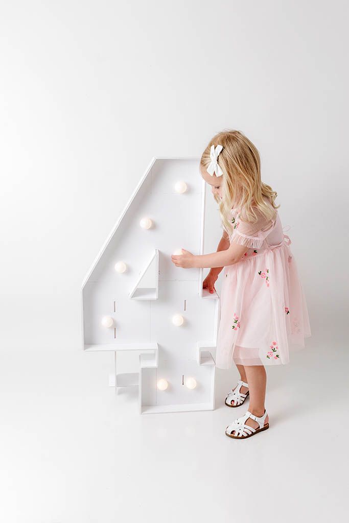 A little girl in a pink tulle dress touches a large illuminated number 4 prop for her 4th birthday photo.