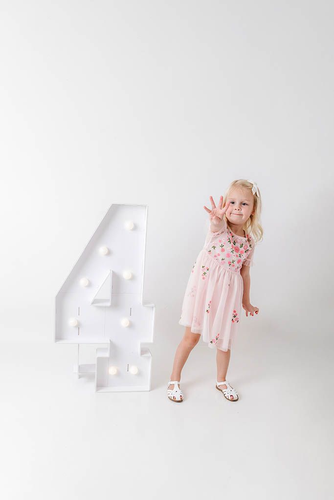 A young blonde girl in a pink floral dress poses next to a large illuminated number 4 on a white background.