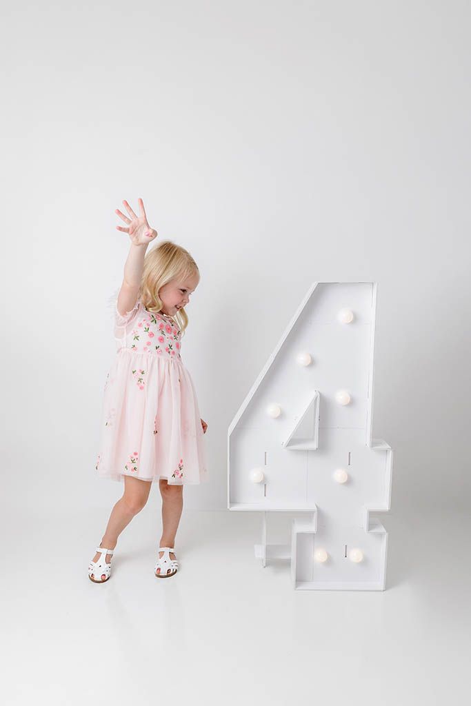 Blonde toddler girl in pink floral dress holding up four fingers beside a large marquee number 4.