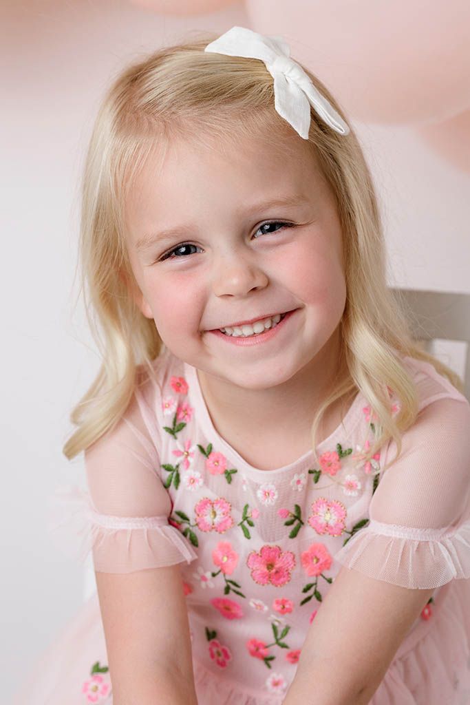 Close-up of happy blonde little girl in pink embroidered floral dress with white bow.