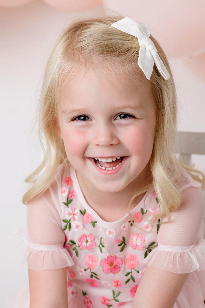 Smiling blonde little girl in pink floral dress with white bow hair clip against soft pink background.