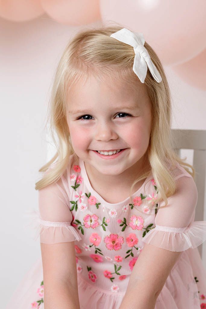 Happy blonde toddler girl wearing pink floral dress and white bow smiling while seated near pink balloons.