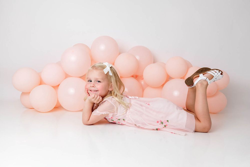 Young girl in pink floral dress lying on floor smiling with pink balloon backdrop.