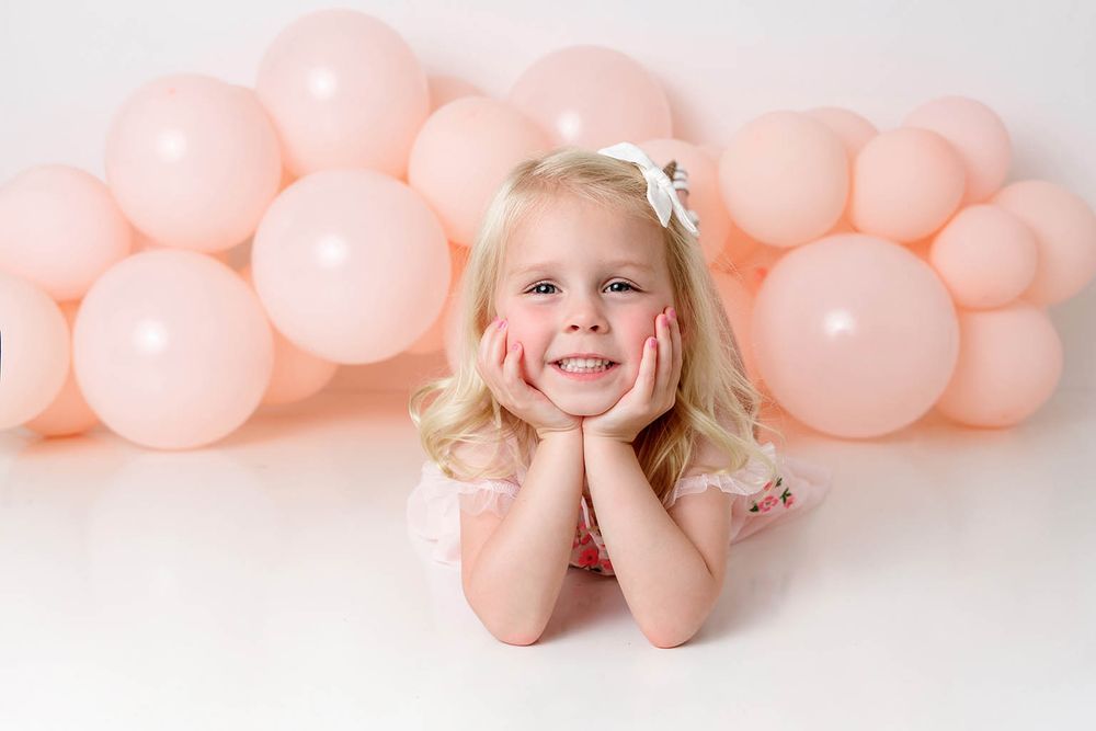 Smiling blonde girl in pink dress posing with hands on cheeks against pink balloons.