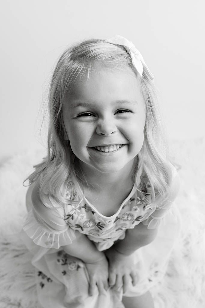 Black and white portrait of joyful blonde toddler girl in floral dress smiling brightly.