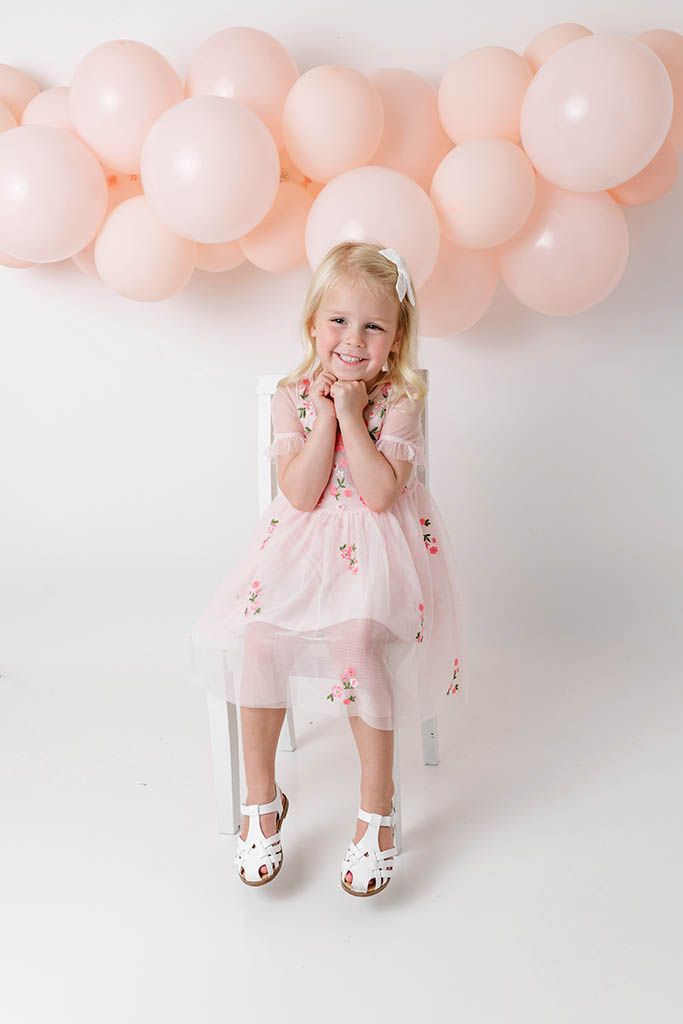 Cute blonde girl in pink embroidered tulle dress sitting and smiling with pink balloons.
