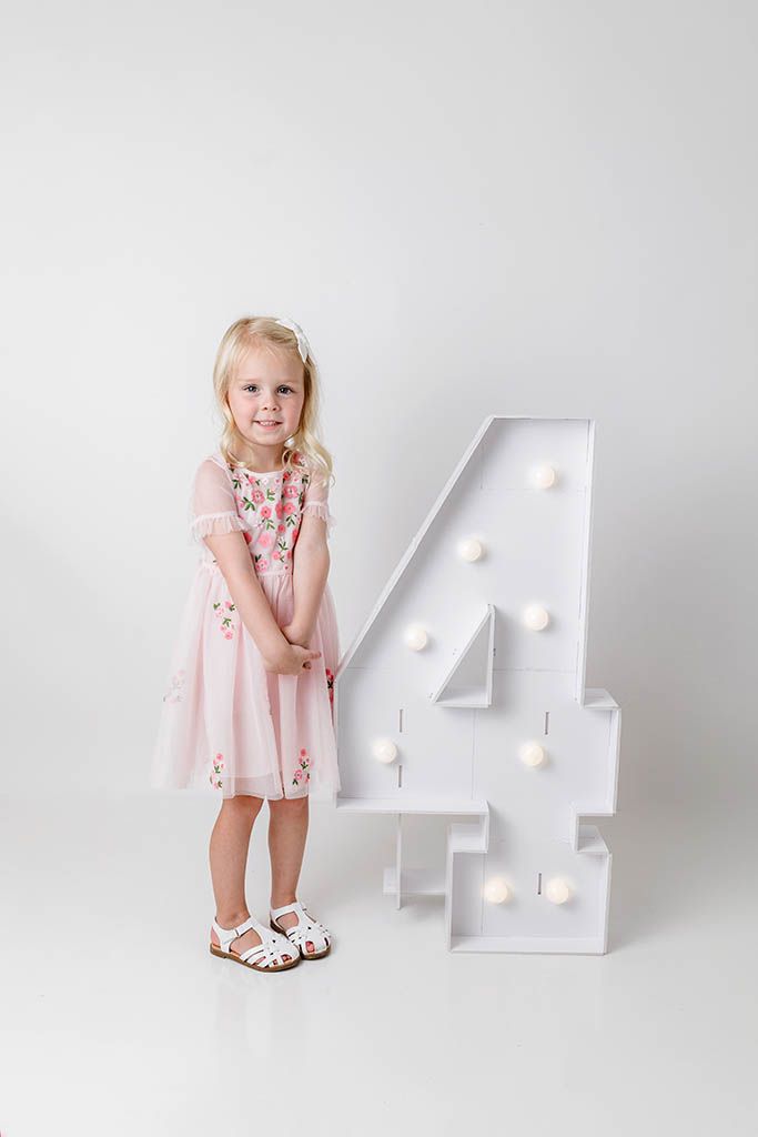 Blonde toddler girl in pink floral dress posing beside illuminated number 4 decoration.