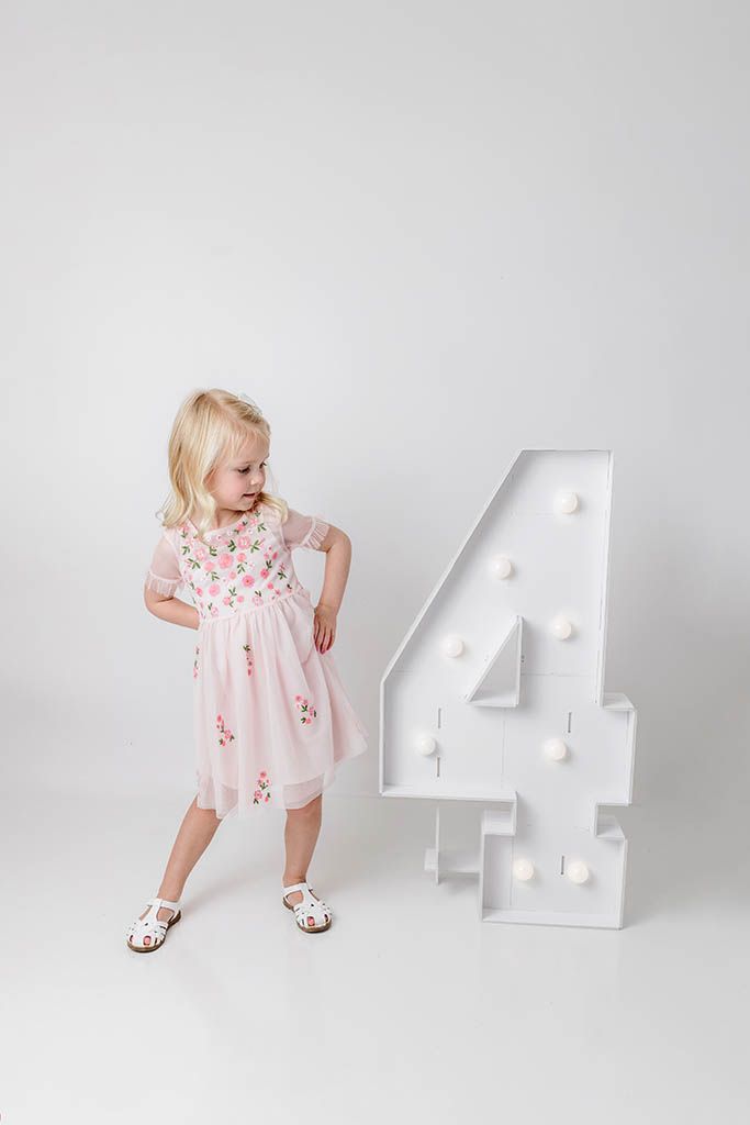 A young blonde girl in a pink floral dress stands next to a large decorative number 4 on a white background.