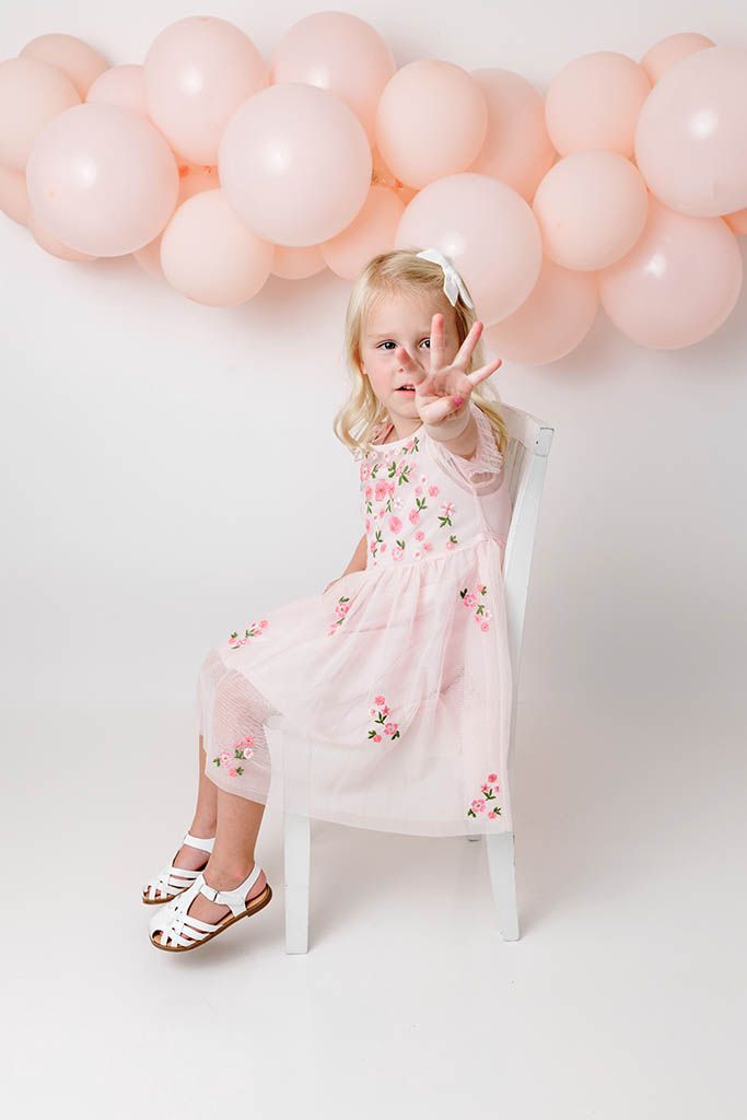 Little blonde girl in pink floral dress sitting on white chair with pink balloons behind.