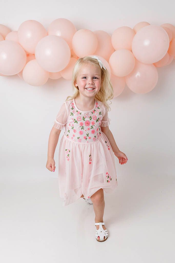 Happy blonde toddler in pink floral dress dancing in front of pink balloon garland.