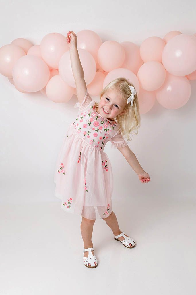 Joyful blonde girl in pink floral dress dancing with arm raised in front of pink balloon garland.
