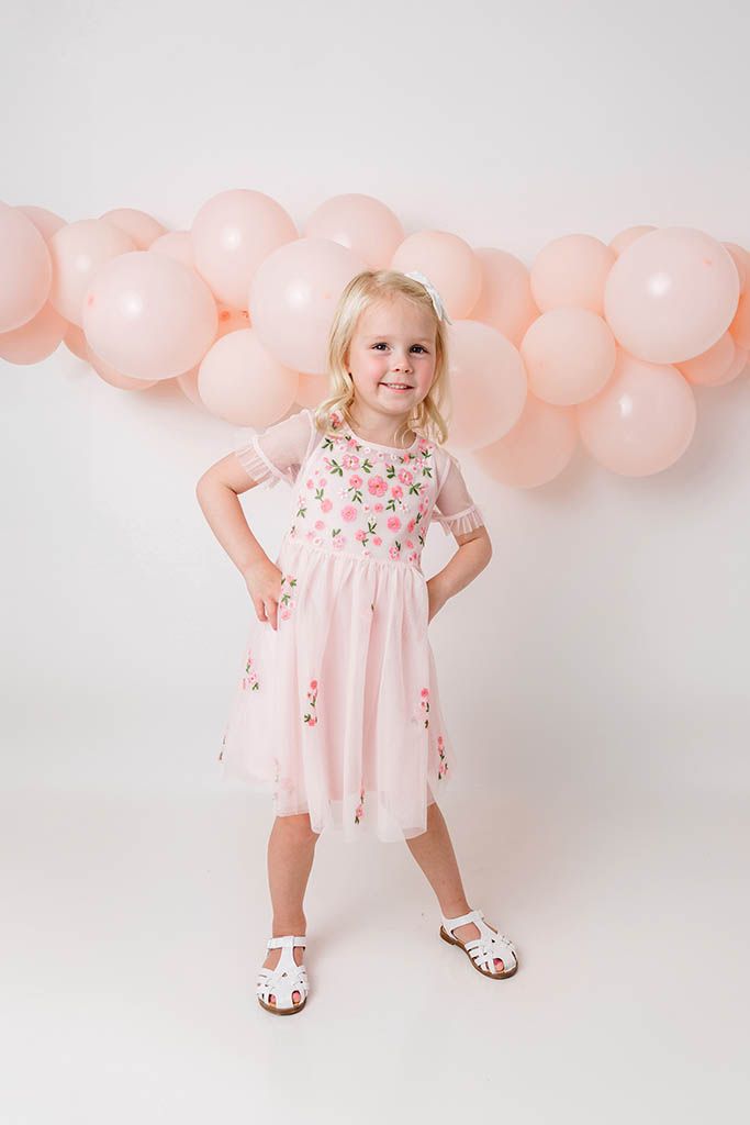 Confident little girl in pink embroidered dress posing with hand on hip by balloons.