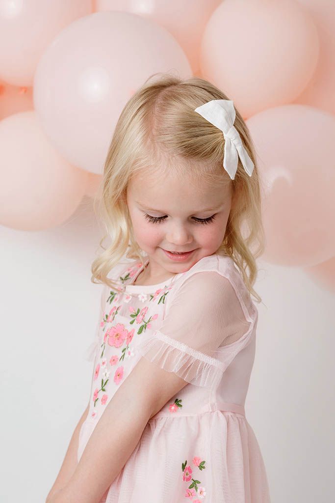 Sweet blonde girl in pink floral dress with white bow looking down and smiling.