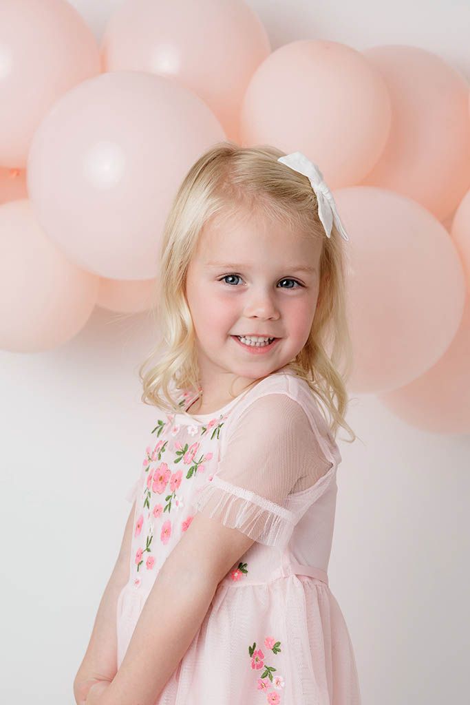 Smiling blonde little girl in pink floral tulle dress posing in front of soft pink balloon backdrop.