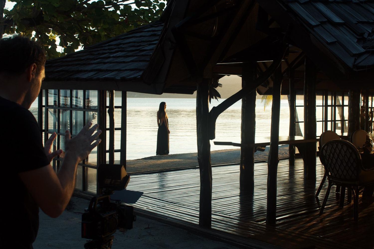 Person photographing a silhouetted figure standing on a tropical waterfront pavilion at sunset.