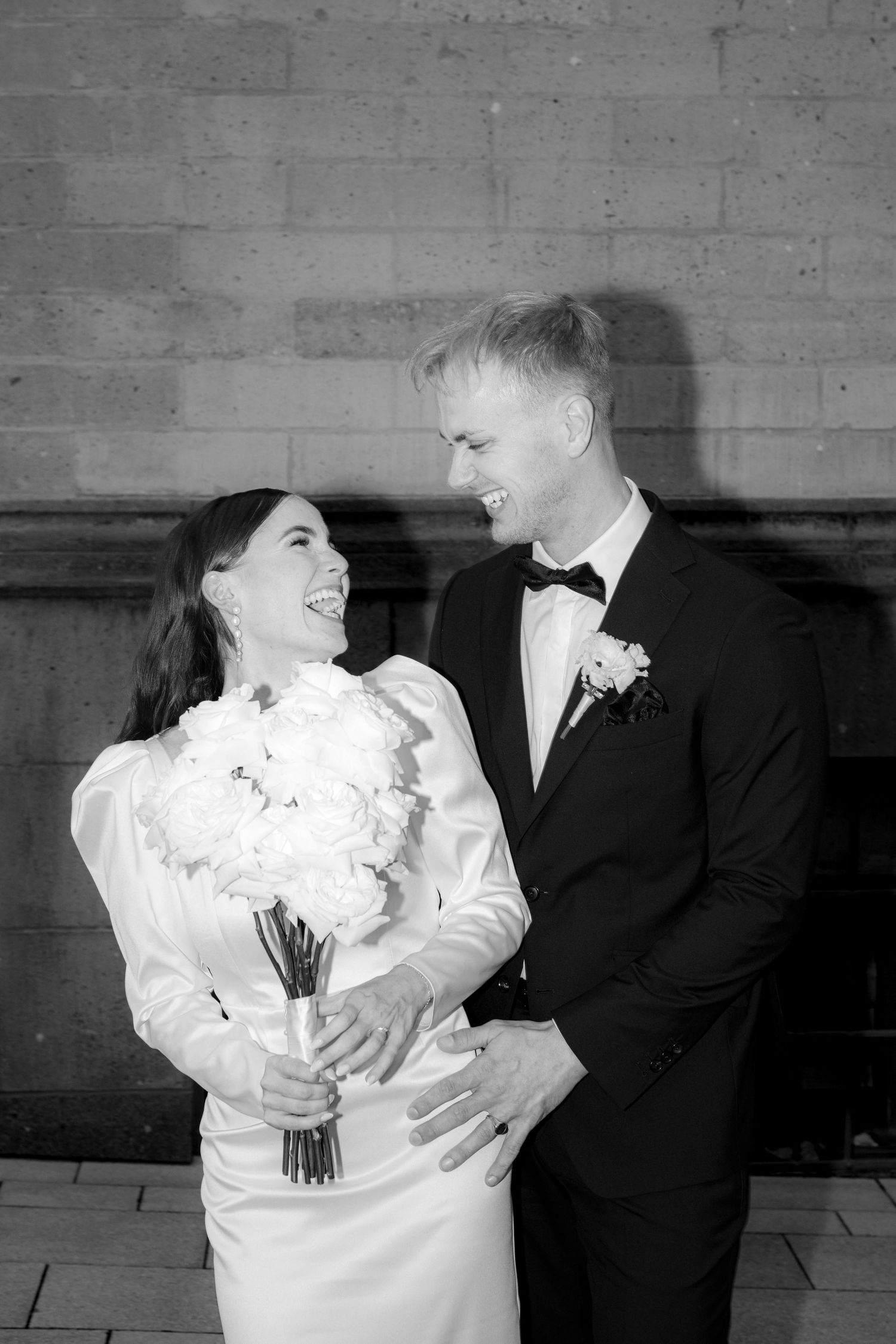 Black and white photo of a smiling bride holding white bouquet and groom in tuxedo at their wedding.