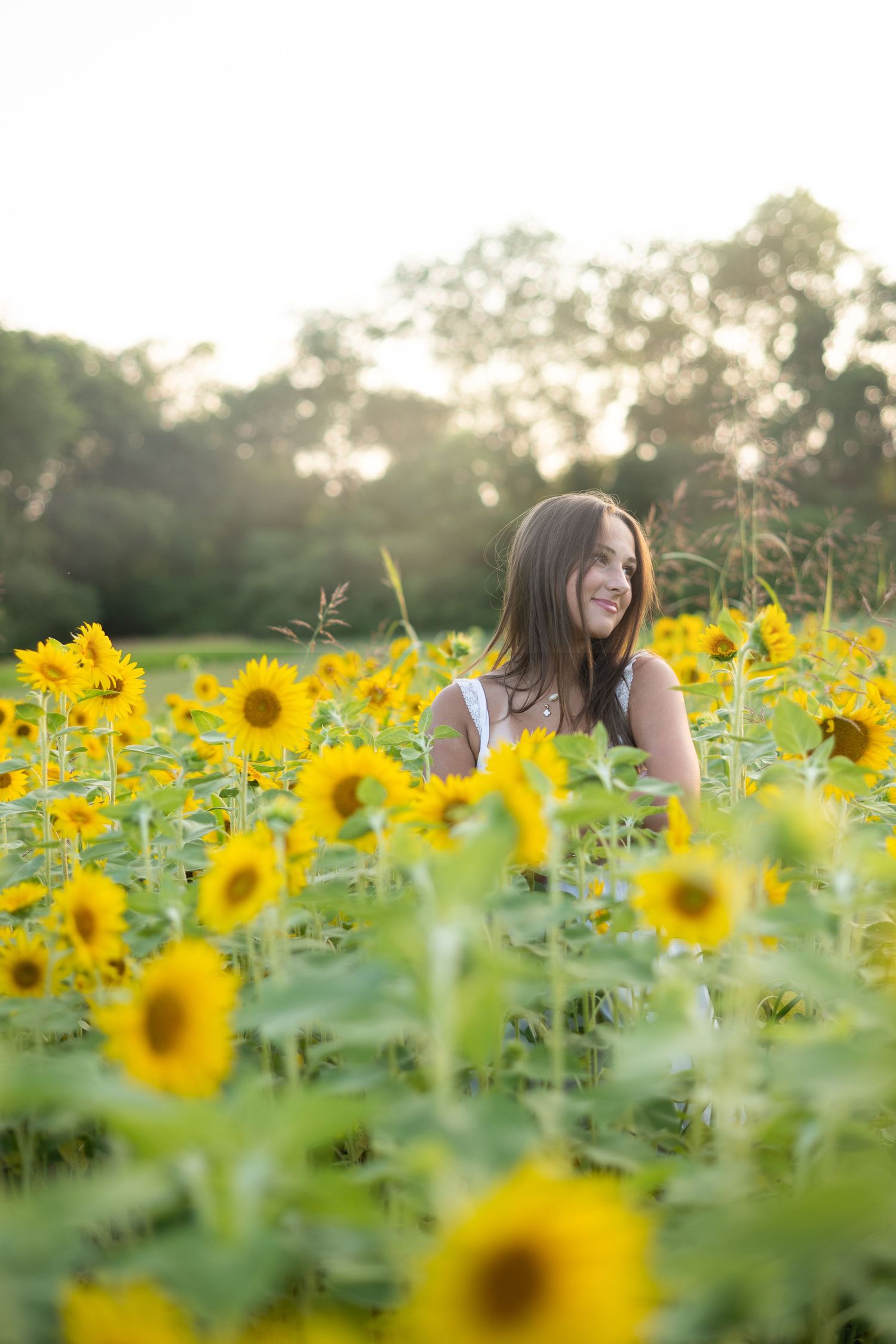 Young woman smiling in a vibrant sunflower field during golden hour, surrounded by bright yellow blooms.