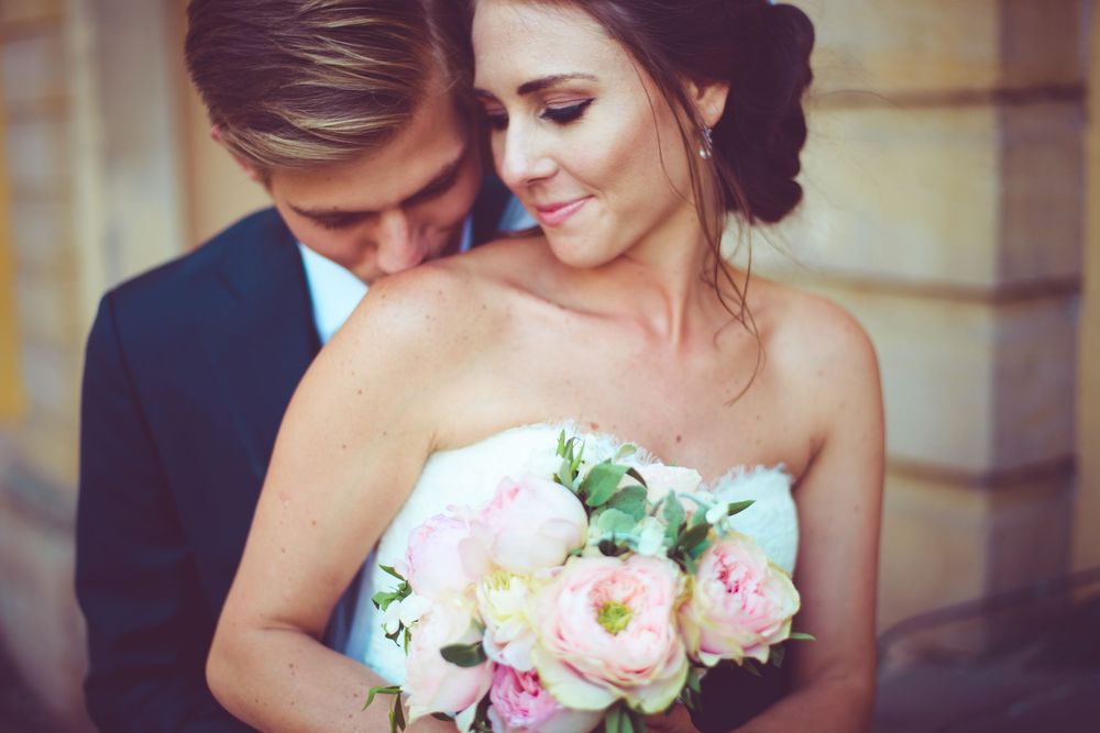 Romantic wedding couple embracing, bride holding pink peony bouquet in strapless gown, groom in navy suit.