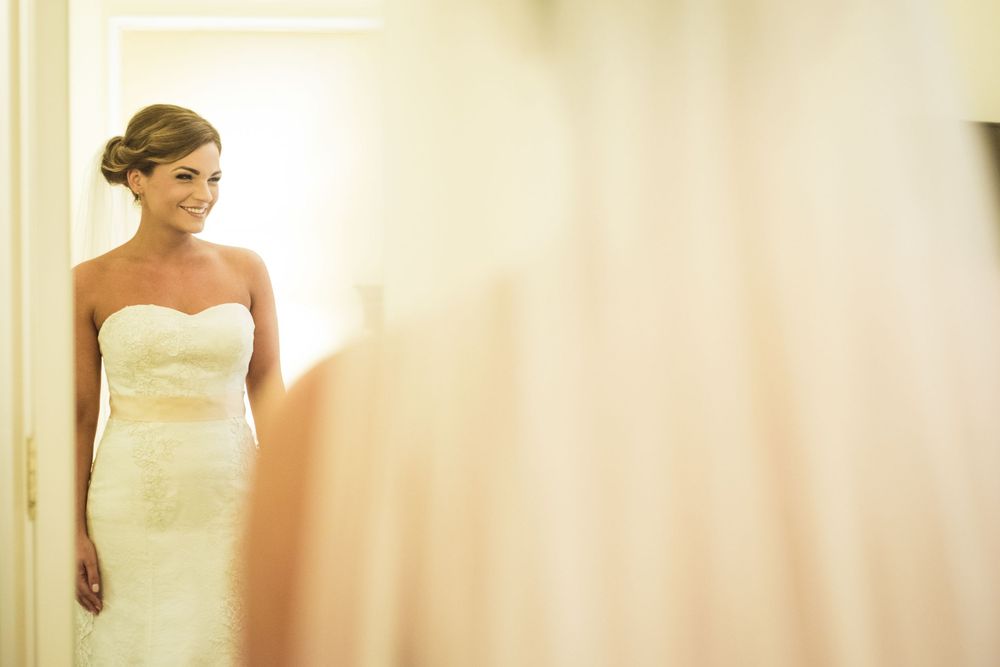 Smiling bride in a strapless white lace wedding gown standing in a softly lit doorway.