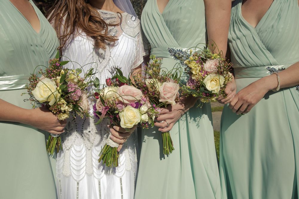Bride in white lace gown and bridesmaids in mint green dresses holding rustic floral bouquets with roses.
