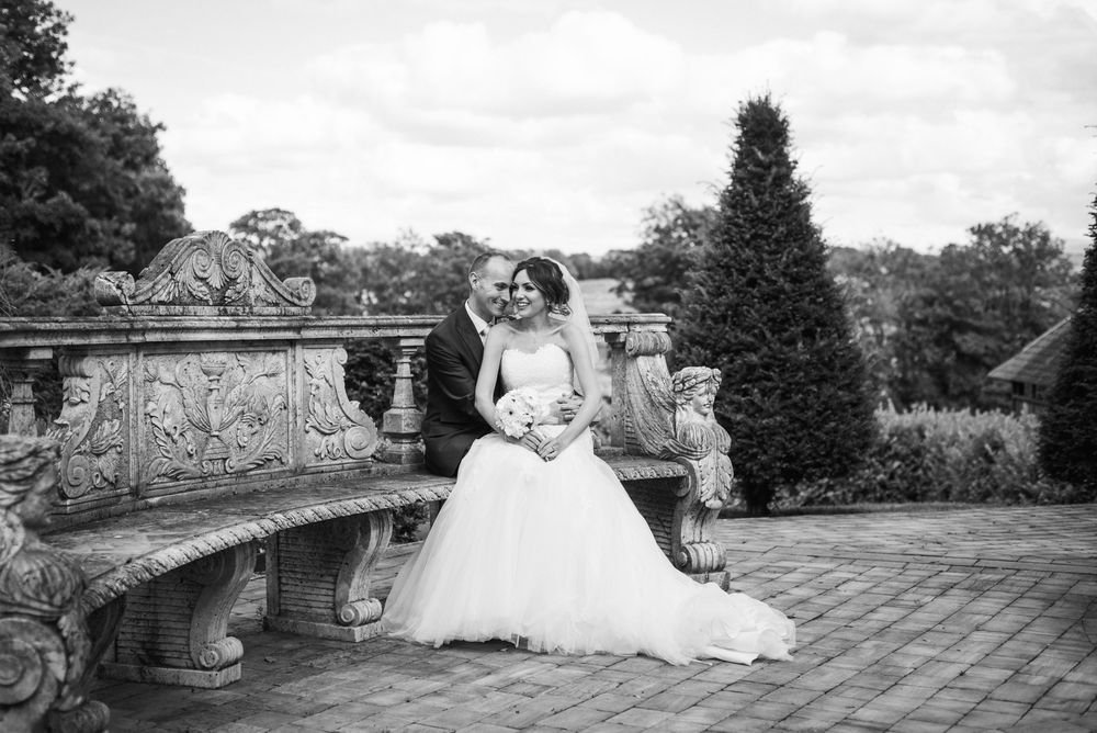Black and white photo of bride and groom posing on ornate stone bench in elegant garden setting.