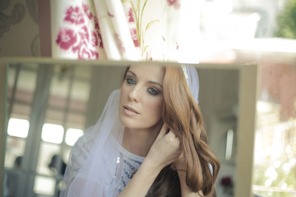 A beautiful bride with long red hair wearing a white veil poses elegantly near a window in soft natural light.