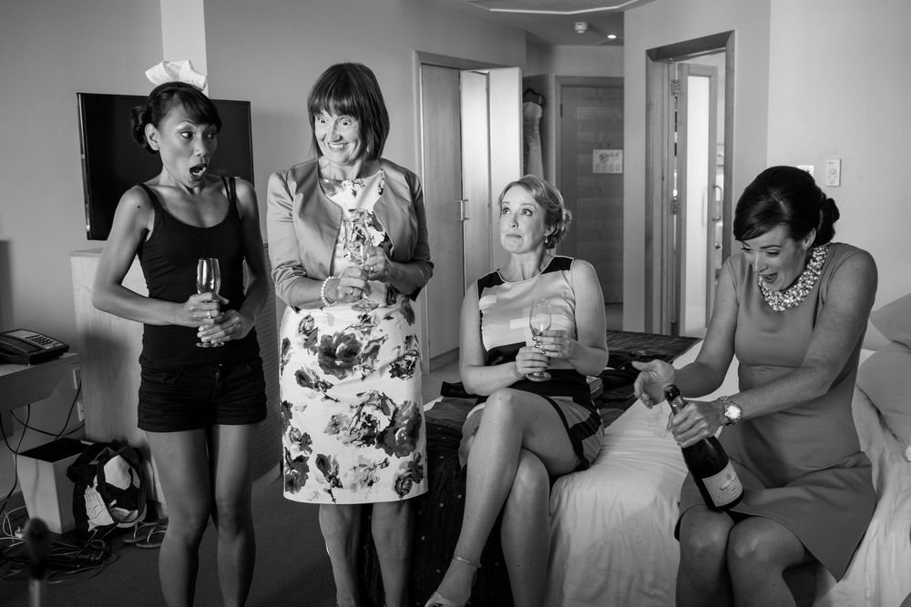 Four women celebrate together in a hotel room, holding champagne glasses in a black and white photo.