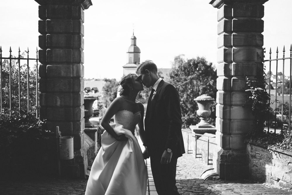 Black and white wedding photo of bride and groom sharing intimate moment under stone archway in elegant garden setting.