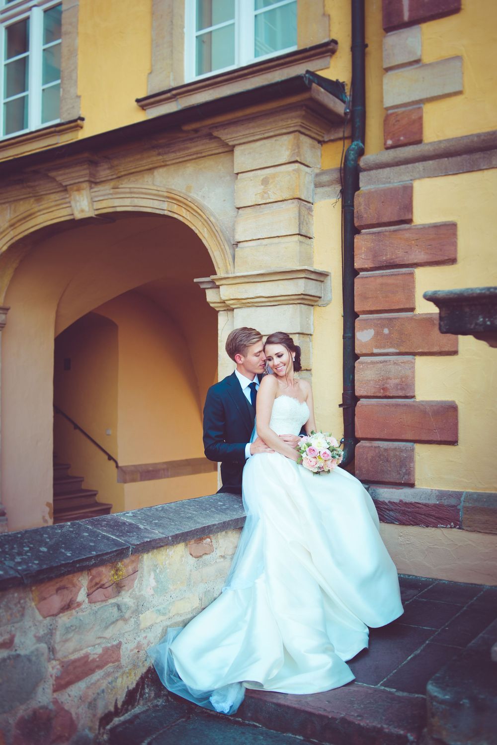 Bride in white gown and groom in suit sitting together on stone steps outside a yellow European building.